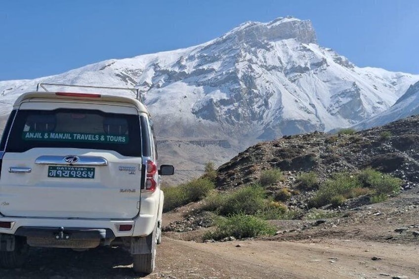 A white SUV with 'Anjil & Manjil Travels & Tours' on the back, parked on a dirt road with a snow-capped mountain in the background. 4