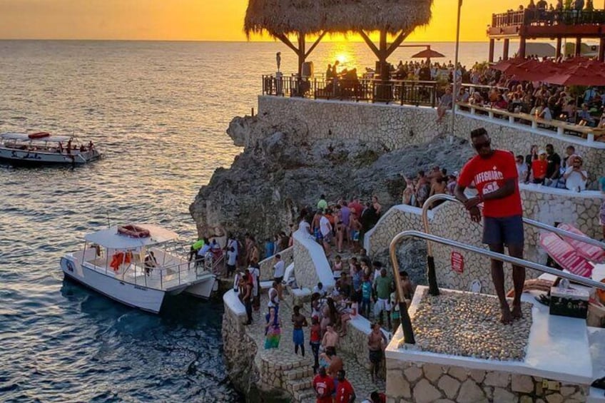 Tramonto su una costa rocciosa con persone su un pontile e una barca nell'acqua. 3