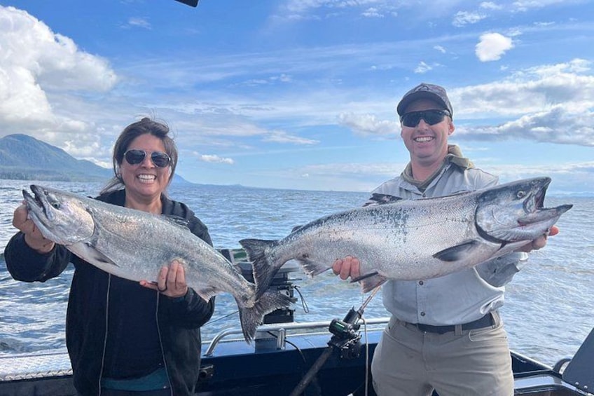 Dos personas sostienen peces grandes en un bote, con montañas al fondo. 4