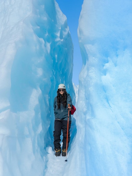 A person standing in a narrow snow crevasse with a red flag, wearing a helmet, sunglasses, and holding a walking stick. 1