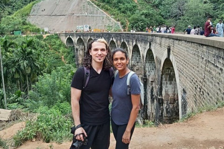 Two people standing in front of a stone bridge with arches, surrounded by greenery. 2
