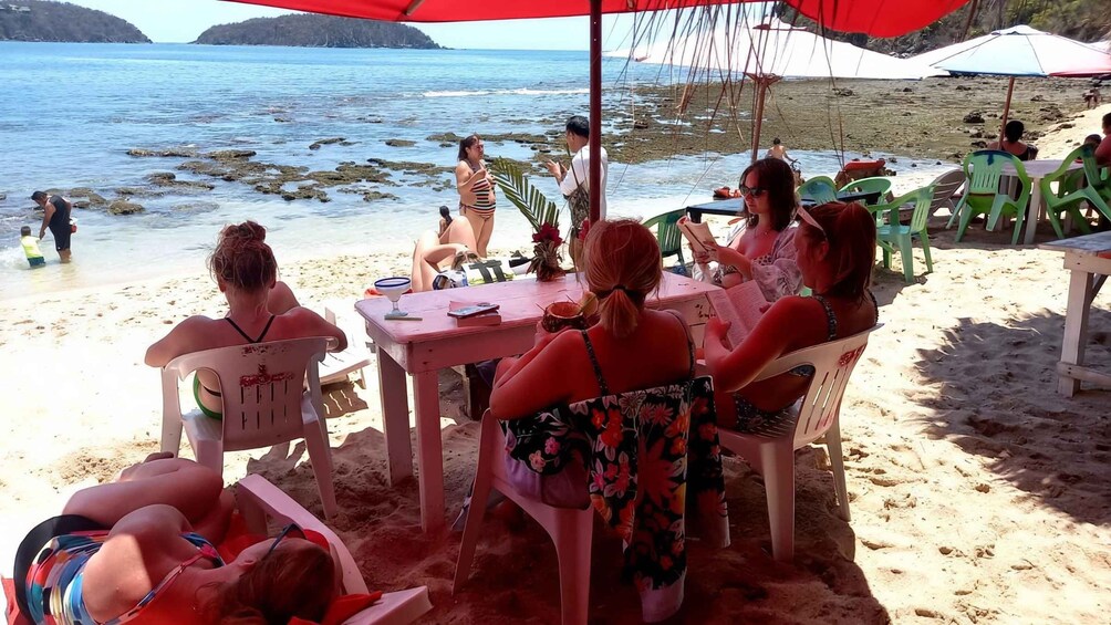 A group of people sitting at a table on a sandy beach with a view of the ocean. 3