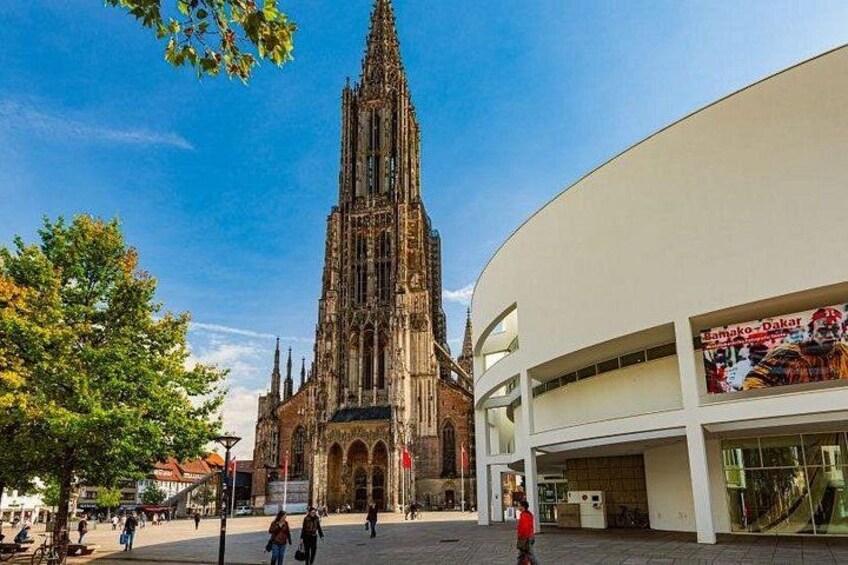 A cathedral with a tall spire, a modern building with a glass facade, and people walking in a square. 4