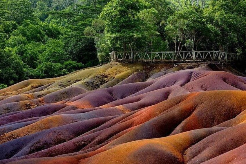 Paisaje con colinas ondulantes cubiertas de tonos vibrantes rojo y naranja, un puente de madera y un bosque verde denso al fondo. 1