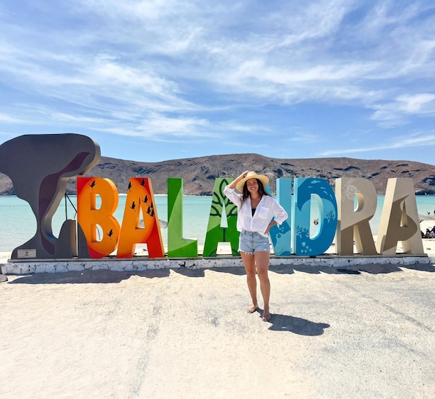 Une personne se tient devant une enseigne colorée sur laquelle on peut lire le mot « BALANDA », avec une plage et des montagnes en arrière-plan. 1