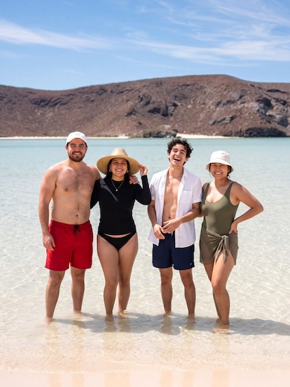 Quatre personnes se tiennent sur une plage, avec une eau cristalline et des montagnes en arrière-plan. 4