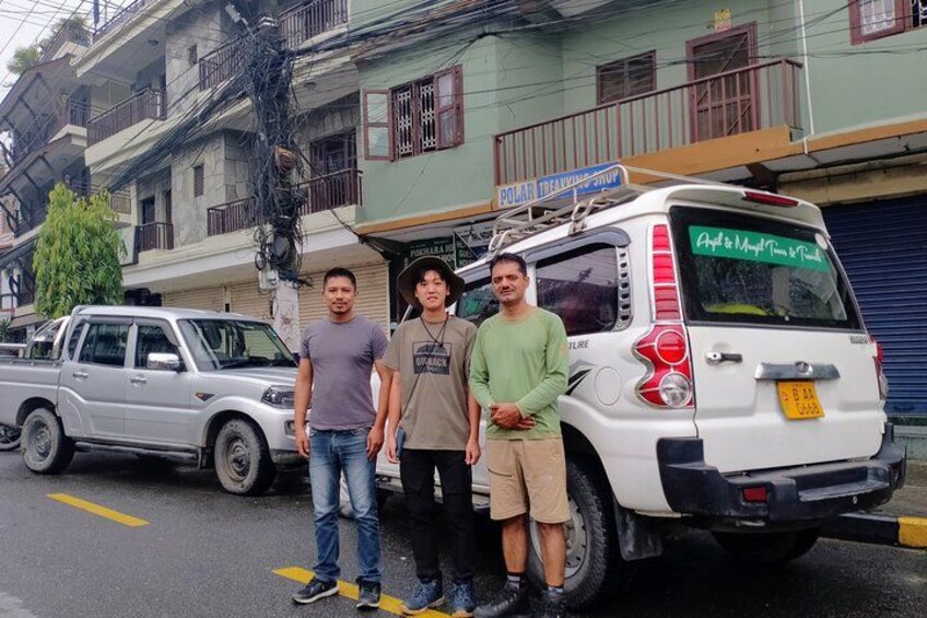 Three people standing next to a white van with a sign on the back, parked on a street with buildings in the background. 5