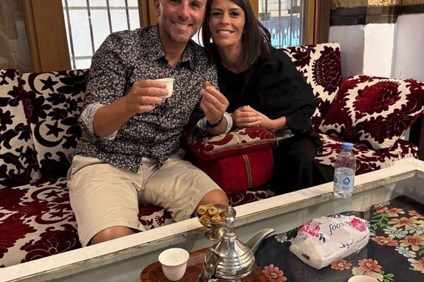 A man and a woman sitting on a patterned sofa, holding cups, with a tray in front of them containing a water bottle, a glass, and a package of tea. 2