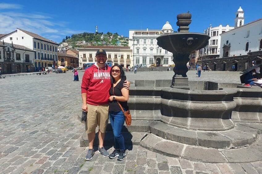 A couple posing in a historic plaza with a fountain, cobblestone pavement, and buildings in the background. 3