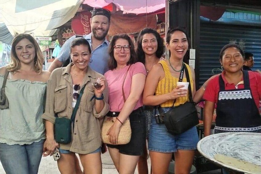 Un grupo de personas en un mercado al aire libre, con varios puestos y letreros al fondo. 1