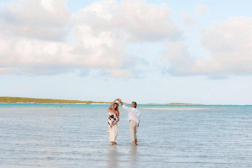 Twee personen staan in ondiep water, één gekleed in een witte jurk en de ander in een wit overhemd, met een rustige zee en een bewolkte lucht op de achtergrond. 3