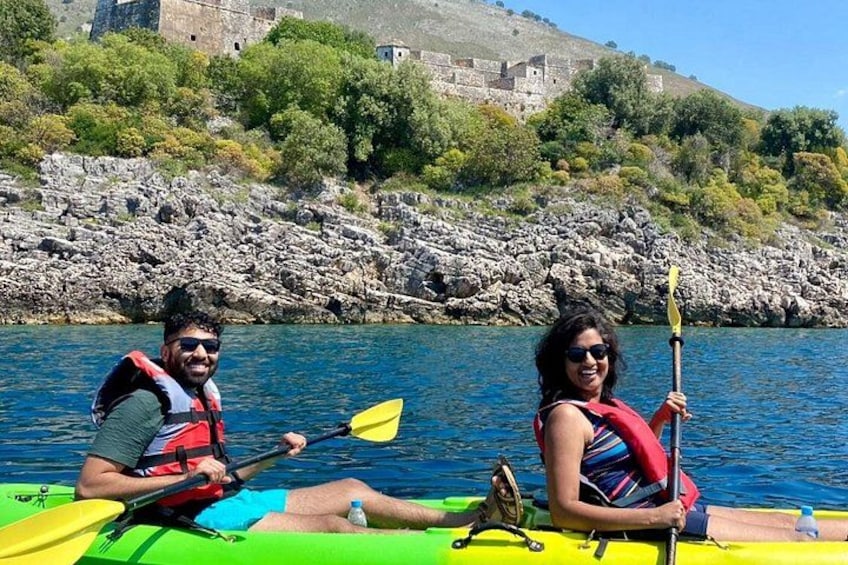 Two people kayaking on a calm river with rocky banks and a historic building in the background. 2