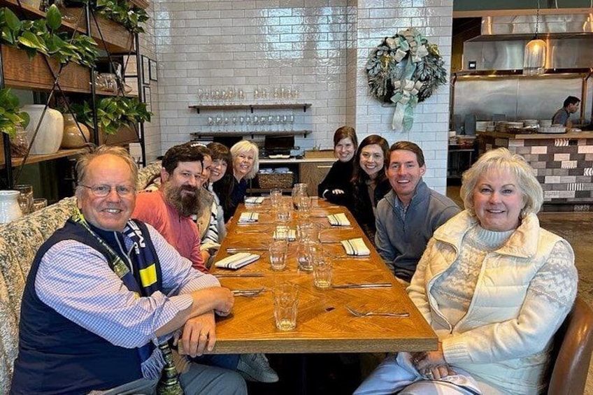 A group of people seated around a wooden table in a restaurant with a brick wall and a kitchen in the background. 2