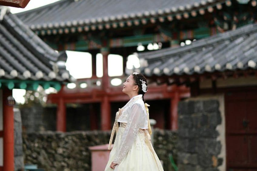 A person in traditional attire standing in front of a traditional Korean building with a tiled roof and stone walls. 1