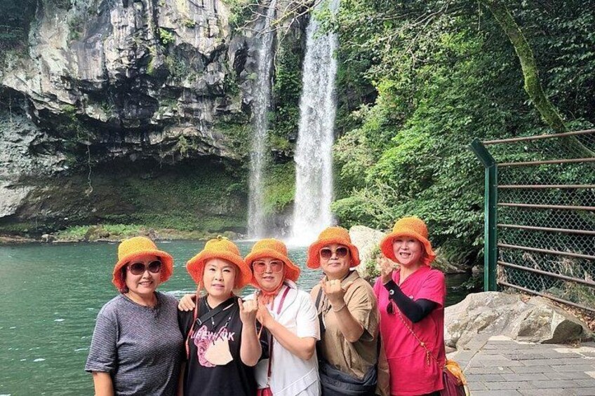 A group of people standing by a waterfall with a rocky cliff and greenery in the background. 2
