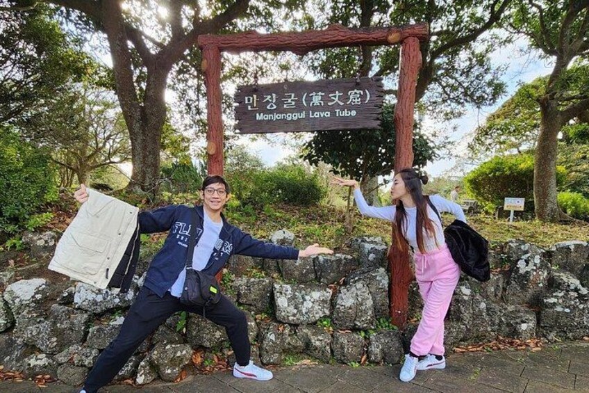 Two people posing in front of a sign with Korean text, a stone wall, and trees in the background. 4