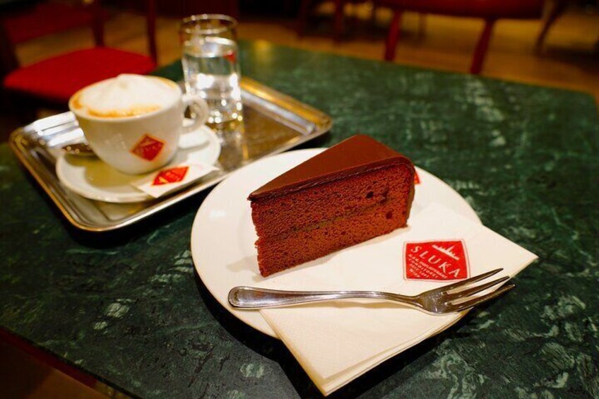 A slice of red cake on a white plate with a fork, a cup of coffee, and a glass of water on a tray. 4