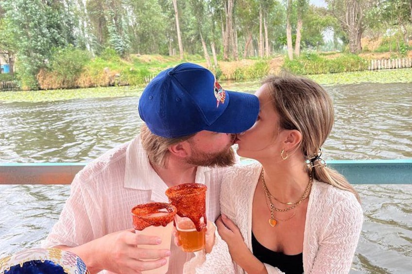 Two people on a boat, holding drinks, with a water body and greenery in the background. 3