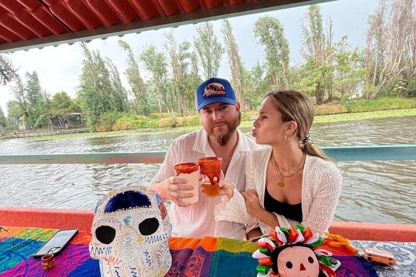 Two people on a boat, holding drinks, with a colorful blanket and a skull decoration on the table. 2