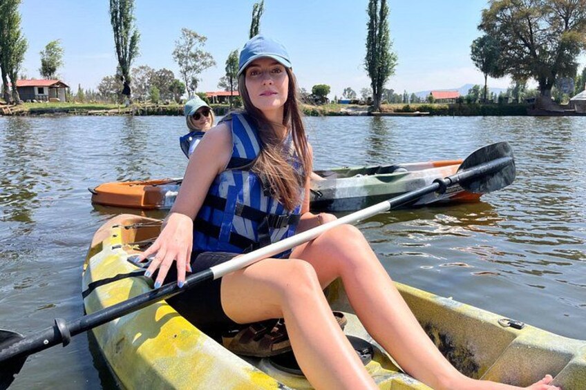 A person in a yellow kayak on a calm body of water with trees and buildings in the background. 1