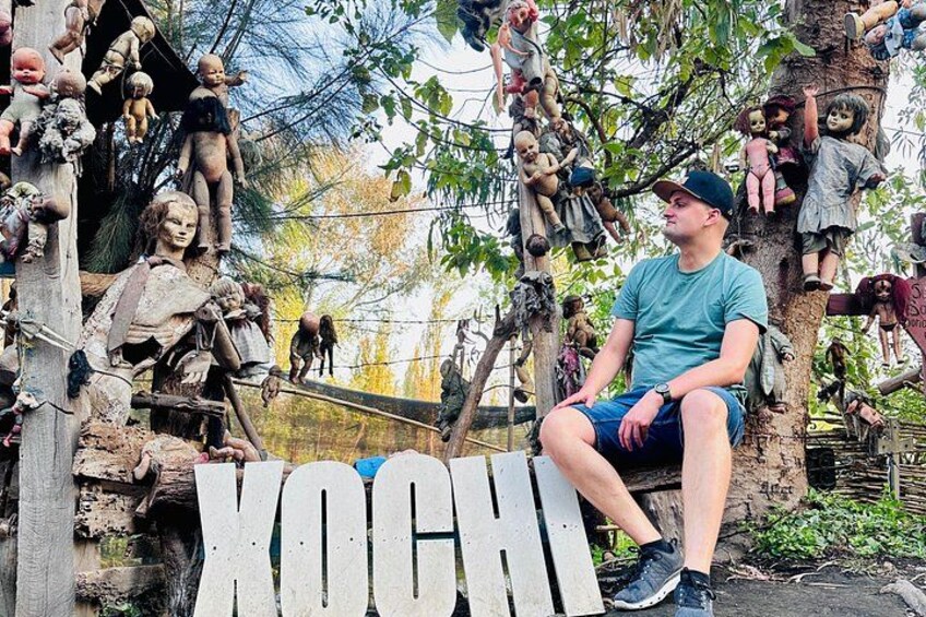 A man sitting on a bench with a tree full of dolls in the background. 3
