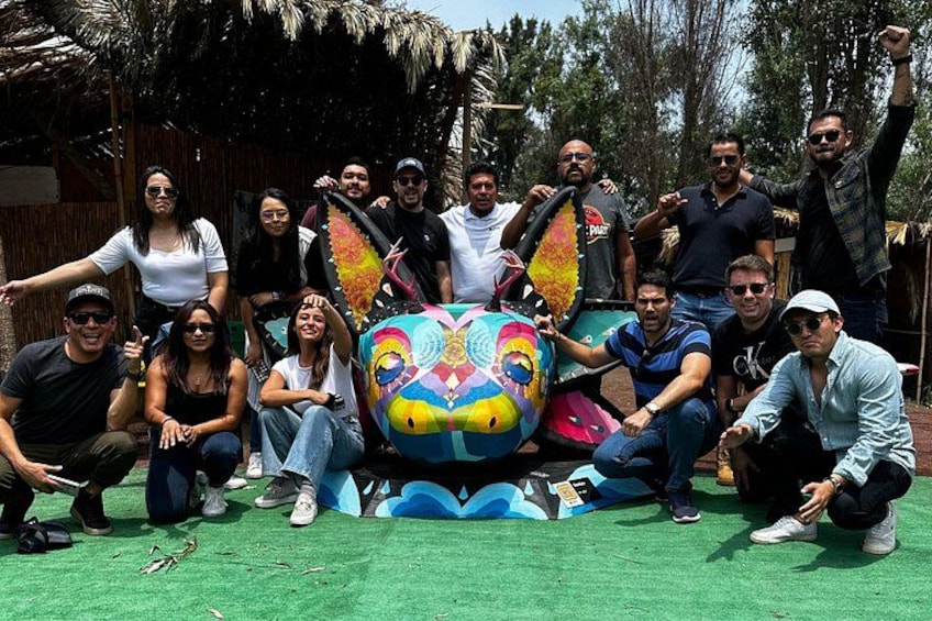 A group of people posing with a colorful, cartoonish face sculpture in a tropical outdoor setting. 2