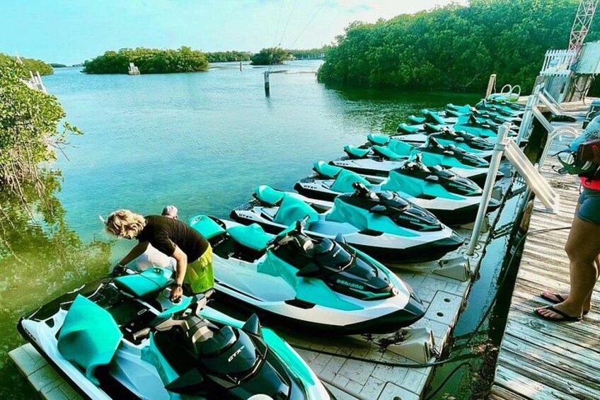 A dock with several jet skis lined up, a person tending to one, and a calm body of water with greenery in the background. 2