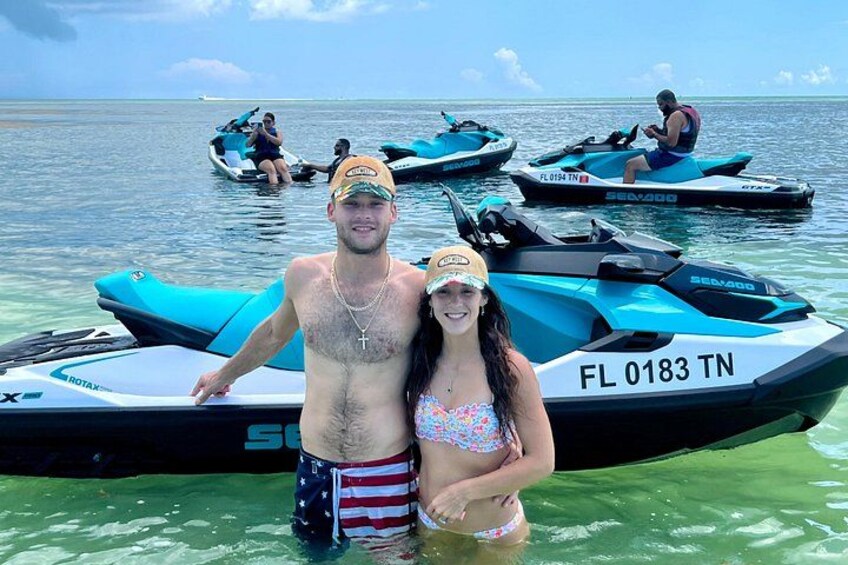 A man and a woman standing in shallow water with jet skis in the background. 5