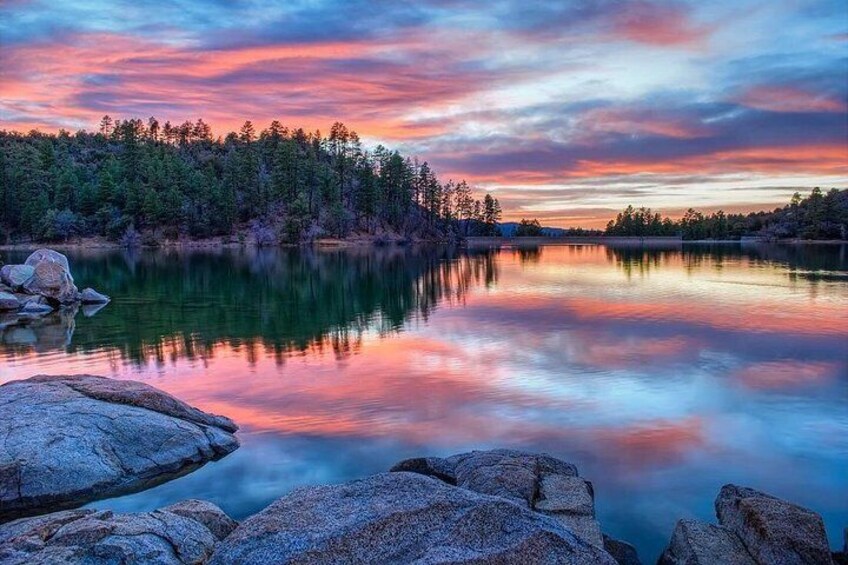 A calm lake with a reflection of trees and rocks under a colorful sunset sky. 1