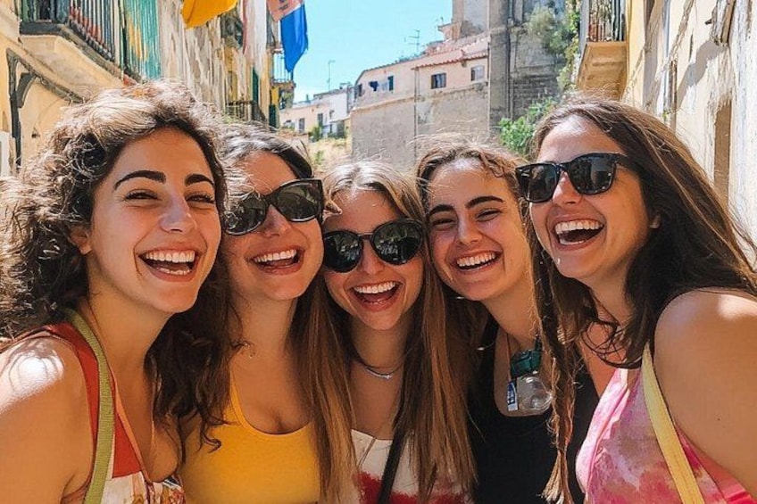 A group of people taking a selfie in a narrow street with buildings and a blue flag in the background. 1