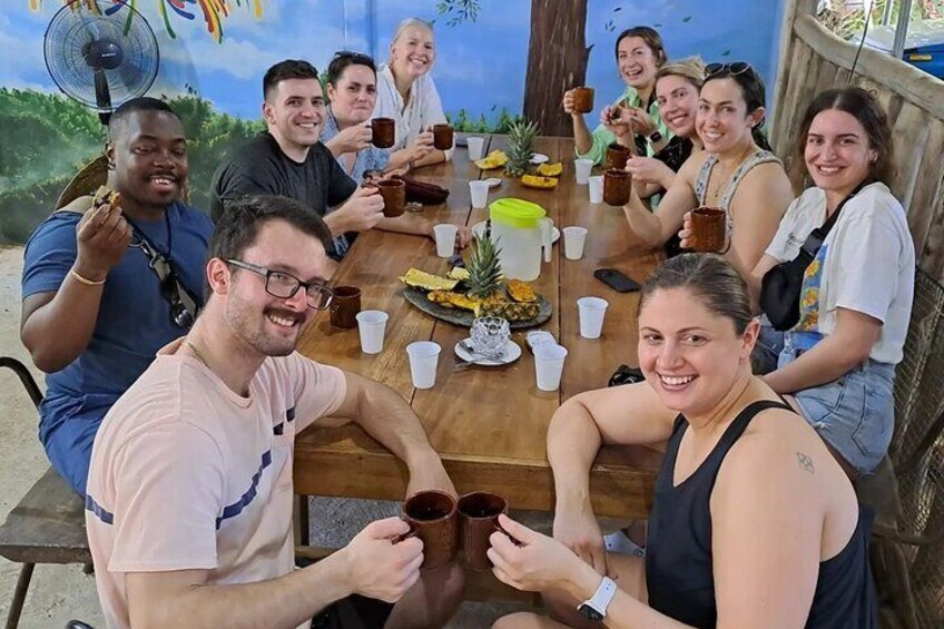 Un groupe de personnes est assis autour d’une table en bois, devant des boissons et de la nourriture, dans un cadre extérieur. Une fresque orne le mur au fond. 2