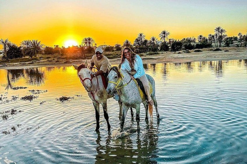 Two people riding horses in shallow water with a sunset in the background. 5