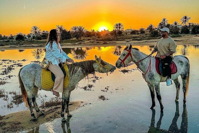 Two people on horses by a reflective water body during sunset. 1