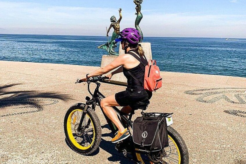 Una persona montando en bicicleta, con una cesta y una mochila, cerca de una escultura y el océano. 2