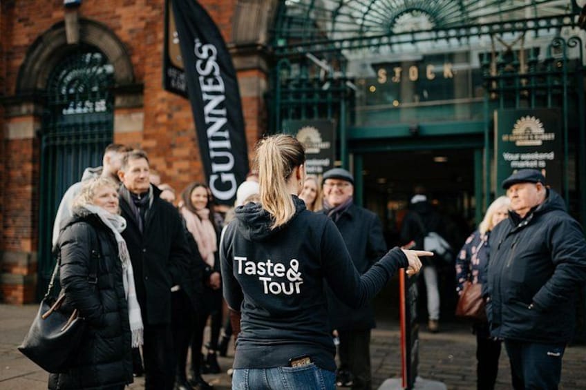 Una persona con una chaqueta negra que tiene 'Taste & Tour' en la espalda, de pie frente a un edificio con un letrero de 'GUINNESS'. 1