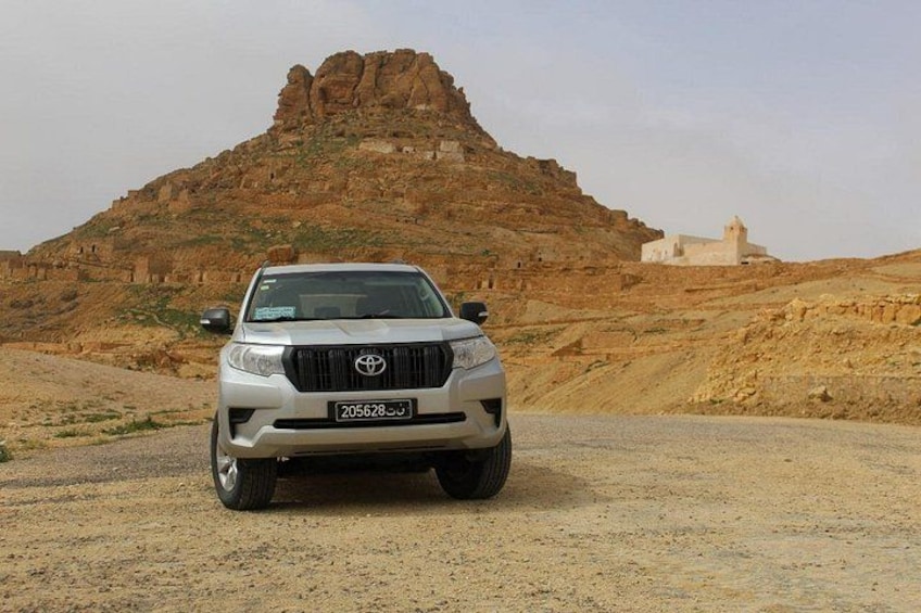 A white Toyota pickup truck parked on a dirt road with a rocky hill and a white building in the background. 1