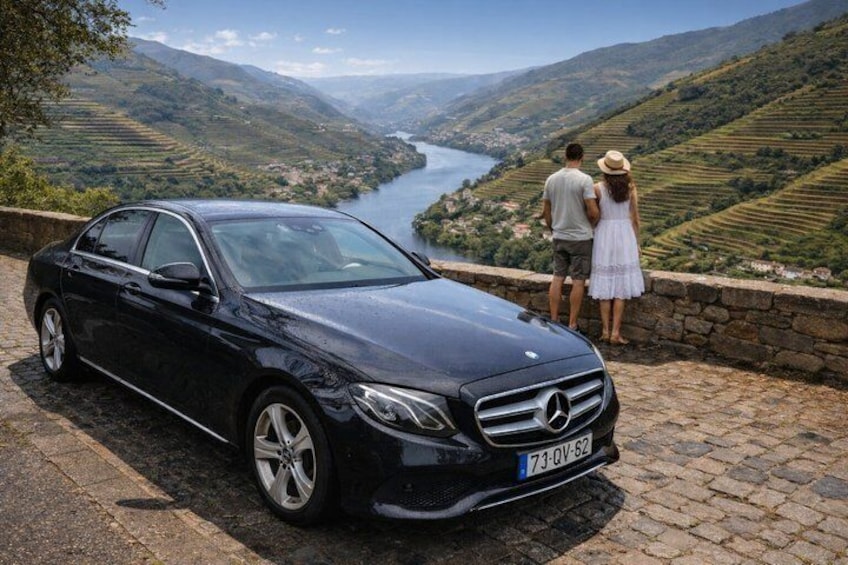 Un coche negro estacionado en un camino adoquinado con vistas panorámicas de un río y colinas al fondo. 1
