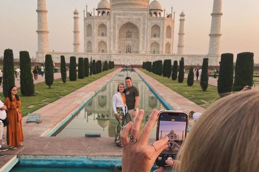 Taj Mahal med et spejlbillede i en bassin, en person tager et billede, og et par står i forgrunden. 3
