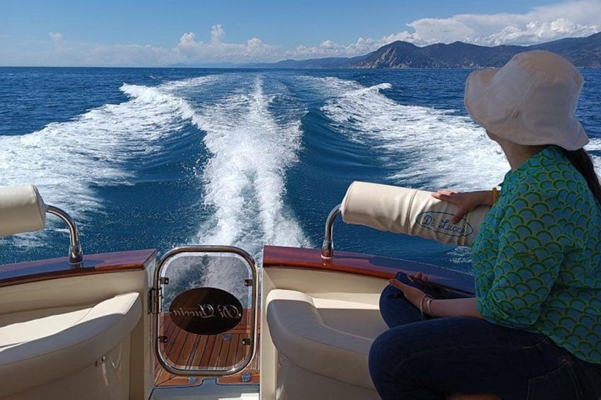 A boat with a woman in a hat, a wake behind it, and a mountainous coastline in the distance. 3