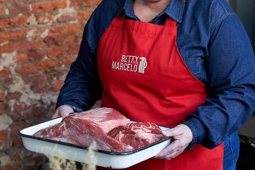 Eine Person hält eine Serviertablett mit rohem Fleisch, trägt eine rote Schürze mit dem Aufdruck 'BETTY AND MARCELO'. 2