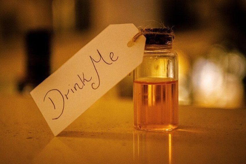 Una pequeña botella de vidrio con una etiqueta que dice 'Drink Me' sobre una superficie reflectante. 5