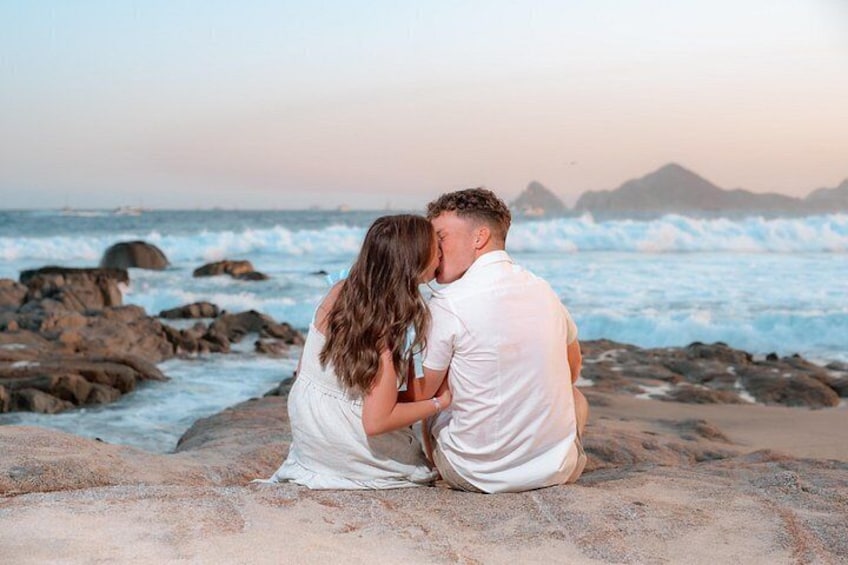 A couple sitting on a rocky beach with the ocean and distant mountains in the background. 5