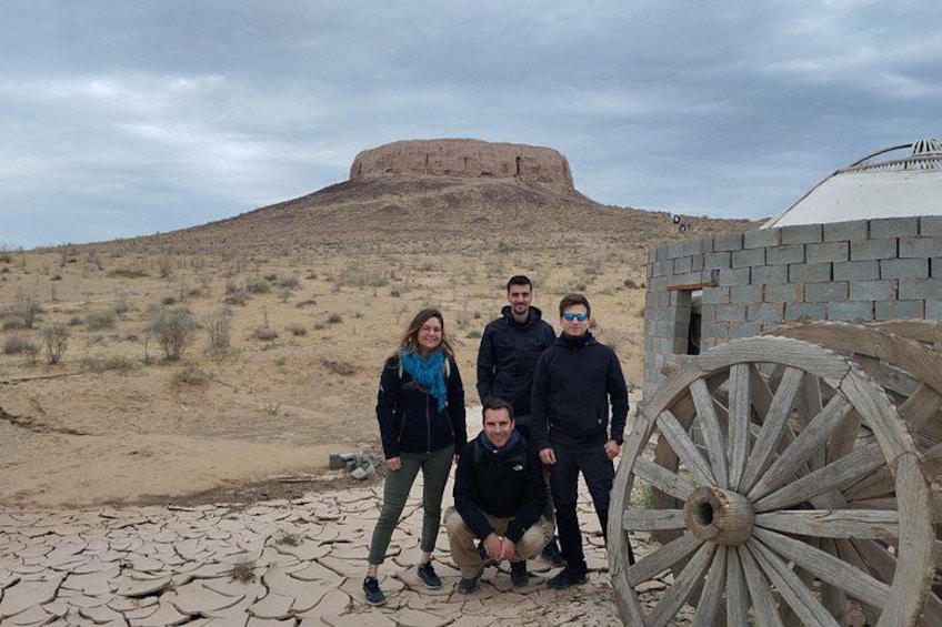 A group of people standing in a desert with a large wooden wheel and a building in the background. 3