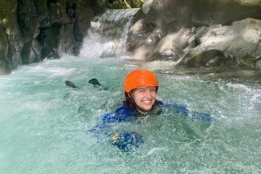 A person in a blue wetsuit and orange helmet in a flowing river with rocks and a waterfall in the background. 1