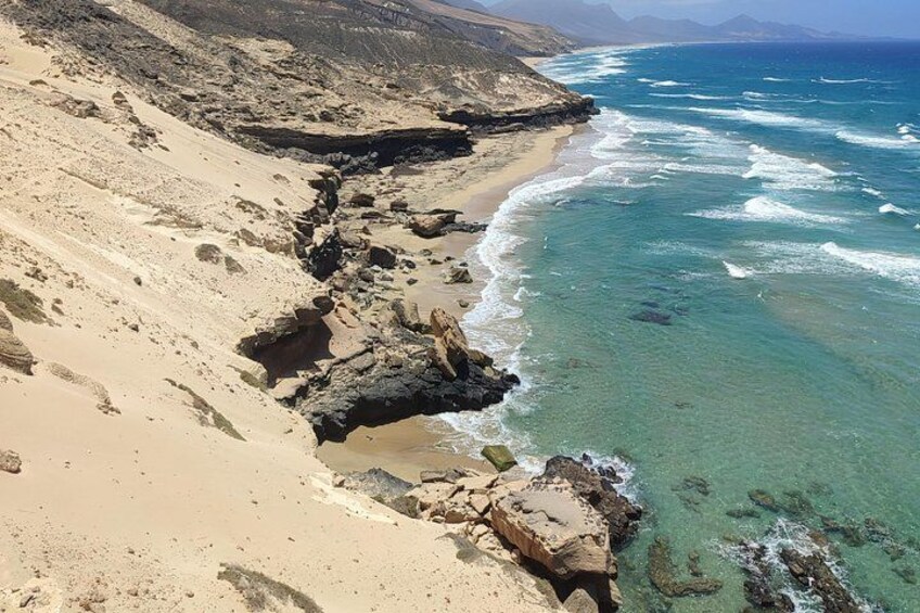 A coastal landscape with a sandy beach, rocky outcrops, and clear blue ocean waves. 2
