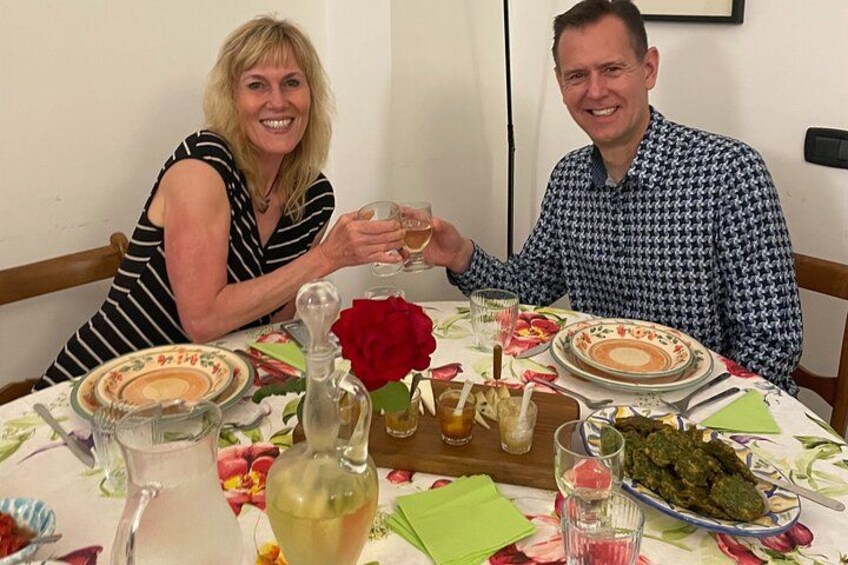 Deux personnes sont assises à une table recouverte d’une nappe à motifs floraux, avec des assiettes, des verres et une rose. 2