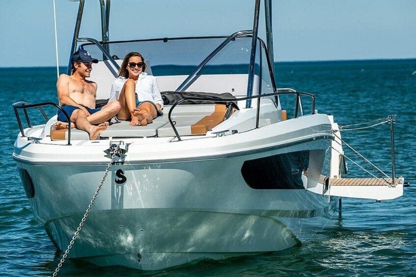 A small white boat with a chain mooring, two people sitting on the deck, and calm blue water in the background. 1