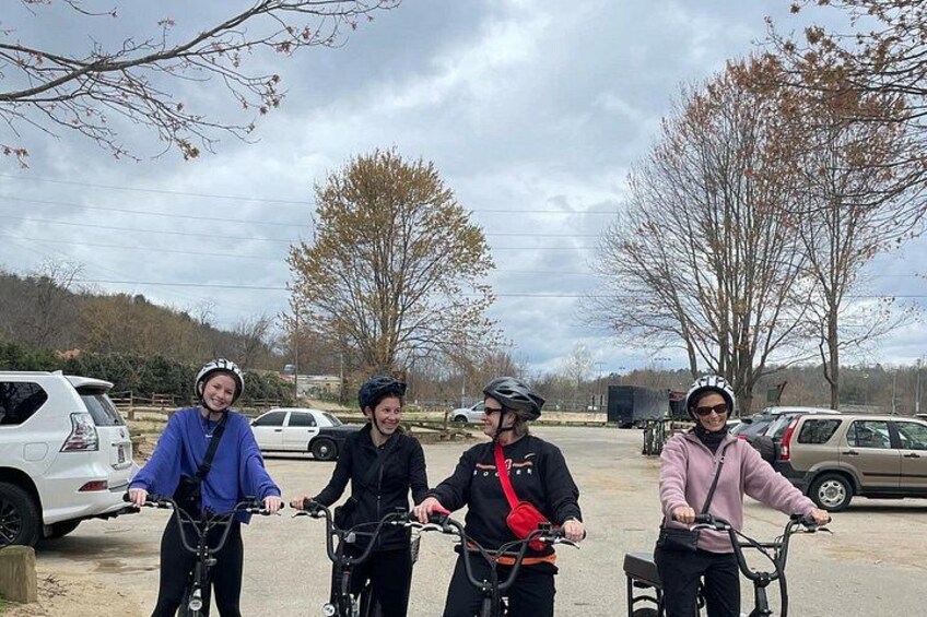 Four people on bicycles in a parking lot with trees and cars in the background. 5