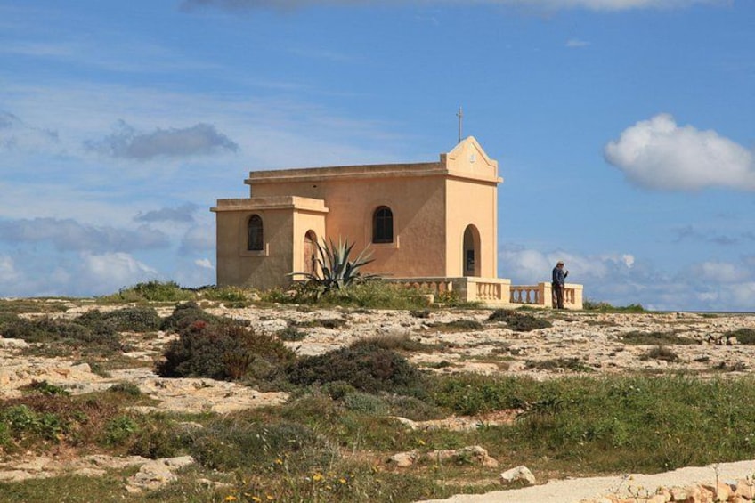 A small, isolated building with a cross on top, surrounded by sparse vegetation and a clear sky. 3