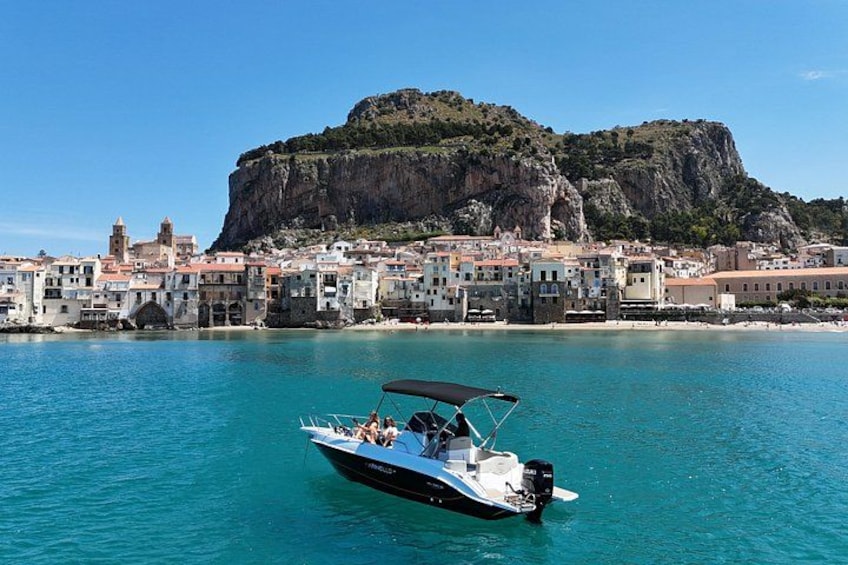 A boat on turquoise water, a coastal town with buildings, and a prominent rocky hill in the background. 1